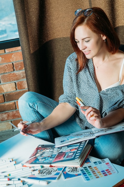 Inspiración artística. Lugar de trabajo. Pintor de mujer pelirroja pensativa mirando paleta con cuadernos de dibujo y suministros alrededor.