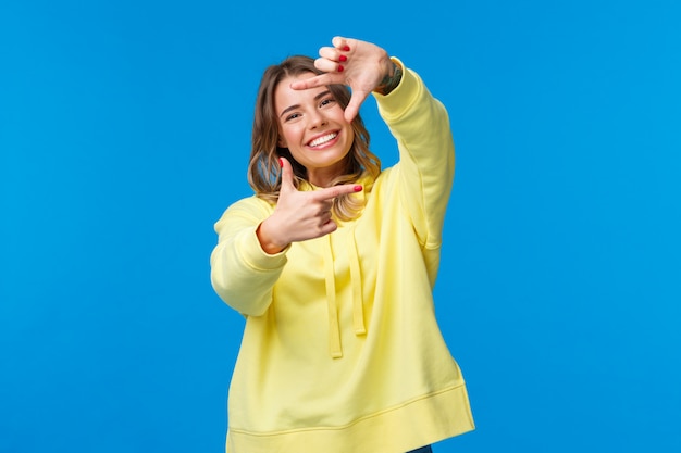 Foto inspiración, estilo de vida y concepto de personas retrato de niña rubia creativa alegre que busca el ángulo perfecto para el disparo, mira la cámara a través del gesto del marco y sonriendo, azul