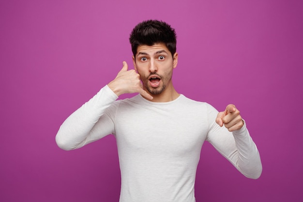 Inspirado joven apuesto mirando y señalando a la cámara haciendo gestos telefónicos con fondo morado