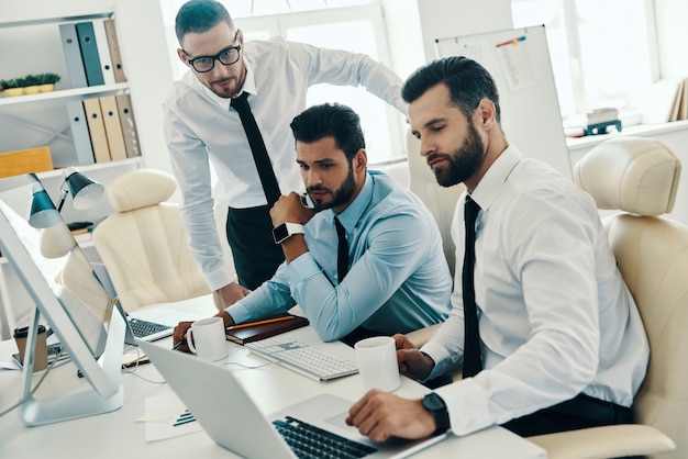 Inspirado para trabalhar duro. Grupo de jovens homens modernos em trajes formais, trabalhando usando computadores enquanto estão sentados no escritório