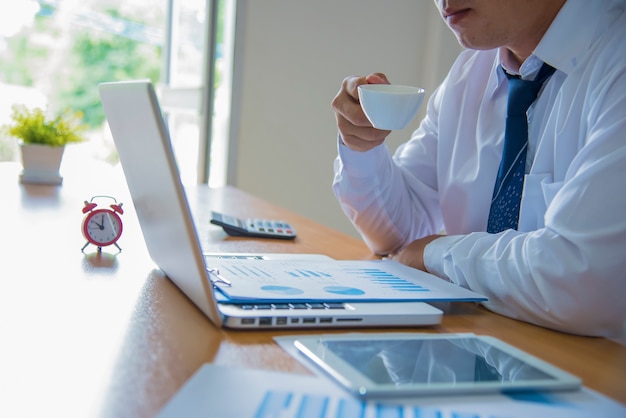 Inspirado con la taza de café fresco. Hombre joven confidente que trabaja en la computadora portátil y que sostiene la taza de café mientras que se sienta en su lugar de funcionamiento en oficina