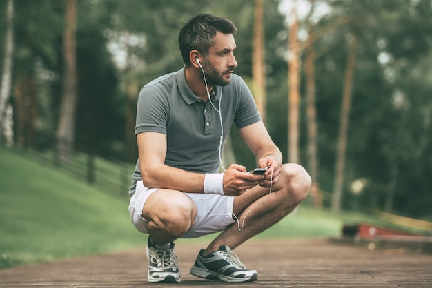 Inspirador con tu música favorita. Hombre joven pensativo en ropa deportiva con auriculares y mirando a otro lado mientras se agacha en el parque