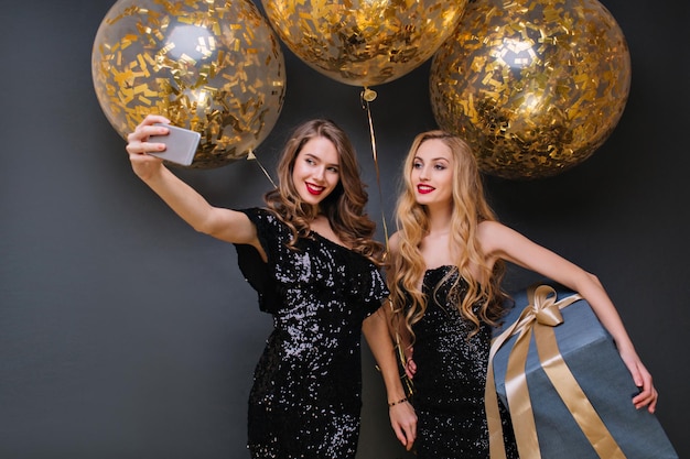 Inspirierte blonde Frau, die ein großes blaues Geschenk hält und auf der Geburtstagsfeier lächelt. Charmantes Mädchen im schwarzen Kleid macht Selfie mit Freund vor goldenen Ballons.