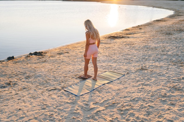 Inspirierte blonde junge Frau tritt auf Sadhu-Brett mit Nägeln am Sandstrand an der Küste im Hintergrund des sanften Sonnenlichts der aufgehenden Sonne am Sommermorgen