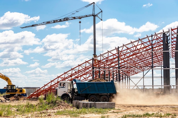 Instalação de treliças e estrutura metálica durante a construção de um edifício industrial ou fábrica O caminhão basculante remove o excesso de solo do canteiro de obras Construção de uma grande oficina de estrutura