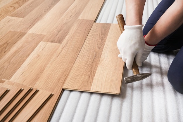 Instalación de piso laminado, detalle de manos de hombre que fijan una loseta con un martillo, sobre una capa base de espuma blanca.