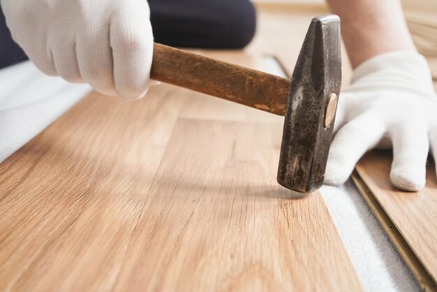 Instalando o piso laminado, detalhes nas mãos do homem em luvas brancas, segurando o martelo sobre a telha de madeira, camada de base branca sob