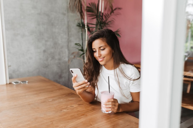 Instantánea de mujer morena con piel bronceada sosteniendo teléfono inteligente y cóctel de fresa