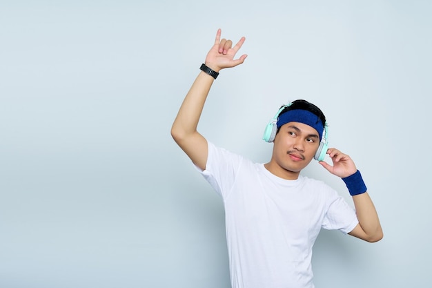 Instructor de entrenador de fitness deportivo de hombre asiático joven loco en diadema azul y camiseta blanca Escuchar música con auriculares Representando el signo de rock de heavy metal aislado sobre fondo blanco