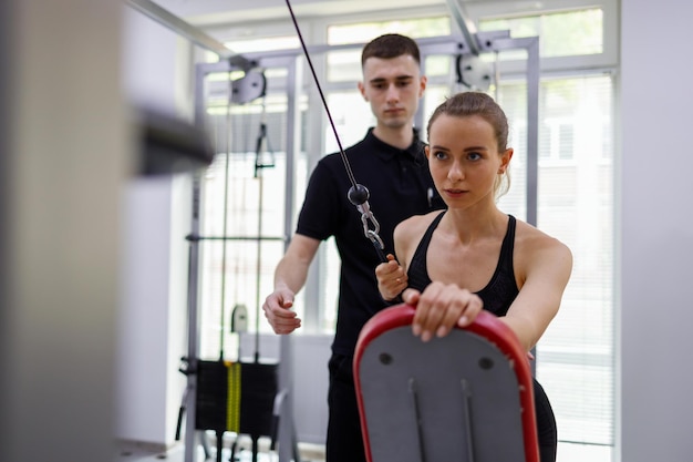 Instructor físico que ayuda a una joven que trabaja en una máquina de ejercicio con cable de tracción