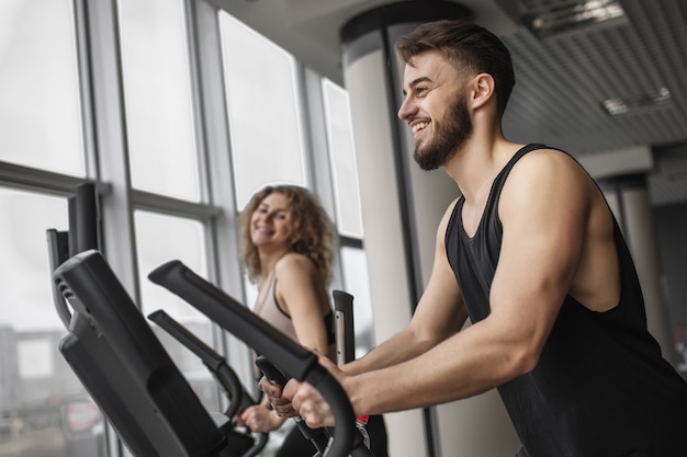 Instructor de fitness guapo está corriendo con su cliente atractivo cómo hacer ejercicio en una bicicleta de ejercicio en el gimnasio