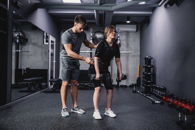 Un instructor de fitness masculino ayuda a las mujeres con un traje especial negro para tecnología EMS mientras realiza ejercicios de estabilidad para la espalda y los brazos en un concepto de gimnasio moderno. Revolución en el entrenamiento, rehabilitación corporal