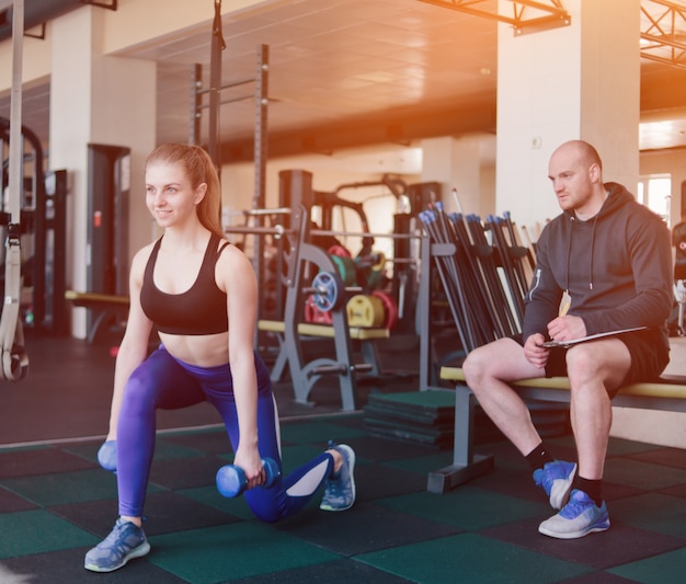 Instructor de fitness supervisa y anota en un cuaderno los resultados del entrenamiento de la joven rubia atlética realizando ejercicios de estocadas con pesas en sus manos en el gimnasio