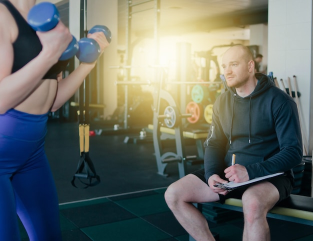 Instructor de fitness supervisa y anota en el cuaderno los resultados del entrenamiento de la joven rubia atlética realizando ejercicios con pesas en sus manos en el gimnasio