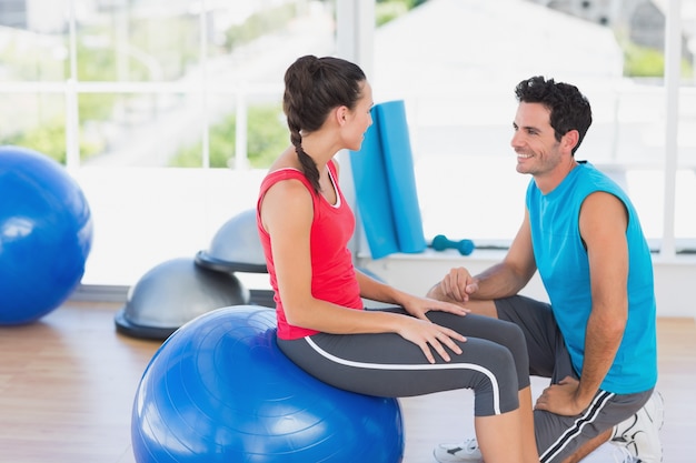 Instructor y mujer con balón de ejercicio en un gimnasio brillante