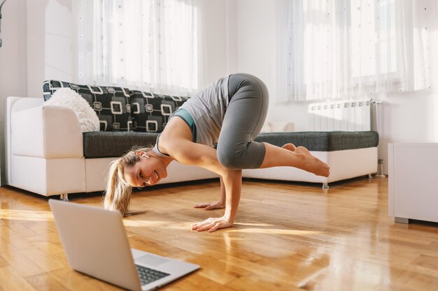 Instructor de yoga sonriente en pose de yoga cuervo con clase en línea sobre portátil