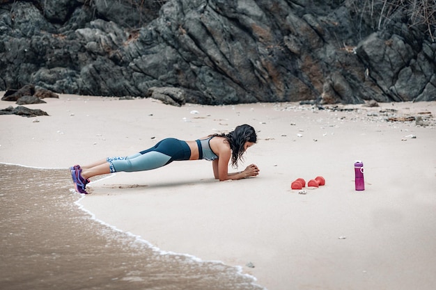 Instructora de fitness deportiva haciendo tablones en la playa de arena cerca del agua, dos campanas rojas y una botella de fitness a su lado.