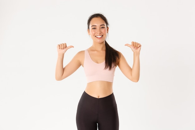 Instructora de fitness femenina asiática sonriente orgullosa y feliz, deportista apuntando a sí misma, ganando objetivo de entrenamiento, fondo blanco.