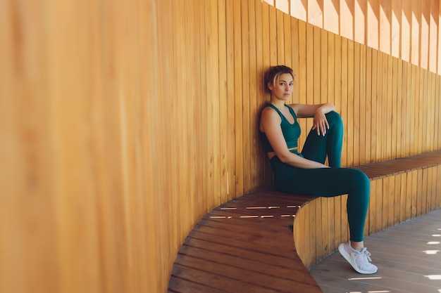 Instructora de fitness joven hermosa da lecciones de video usando la computadora portátil en una terraza de madera en la calle.