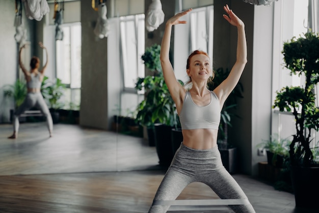 Instructora de fitness pelirroja delgada atlética haciendo sentadillas con banda de resistencia de goma de pilates en las piernas con las manos levantadas sobre la cabeza contra el fondo del espejo en toda la pared en el gimnasio, disfrutando del entrenamiento