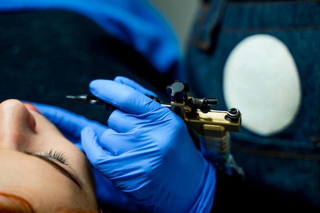 Instrumento negro para maquillaje permanente en manos de cosmetóloga profesional en guantes azules Mujer joven está recibiendo maquillaje de labios permanente Primer plano