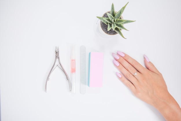 Instrumentos de manicura y mano femenina con uñas rosadas sobre fondo blanco.