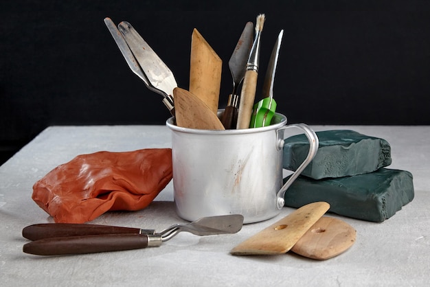 Instrumentos de modelado de plasticina y arcilla en una taza de metal y piezas de arcilla y plasticina en una mesa de hormigón blanco sobre un fondo negro