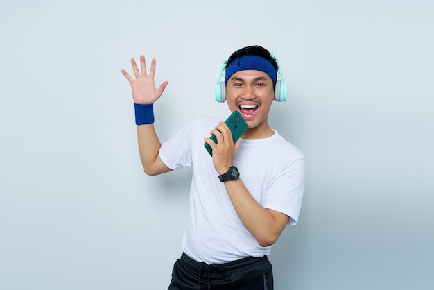Instrutor de instrutor de fitness esportivo jovem asiático alegre na faixa azul e camiseta branca com fones de ouvido ouvindo música cantando música no ditafone no celular sobre fundo branco