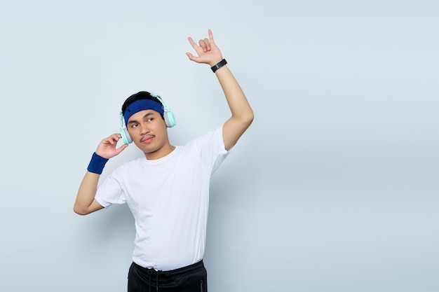 Instrutor de instrutor de fitness esportivo jovem asiático louco em faixa azul e camiseta branca Ouça música com fones de ouvido Representando sinal de rock heavy metal isolado sobre fundo branco