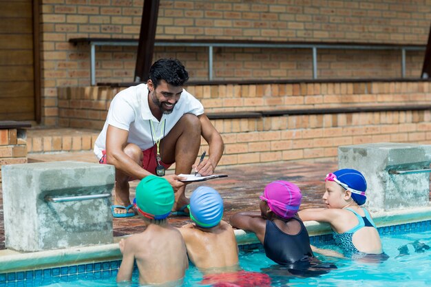 Instrutor escrevendo na prancheta enquanto explica os pequenos nadadores à beira da piscina