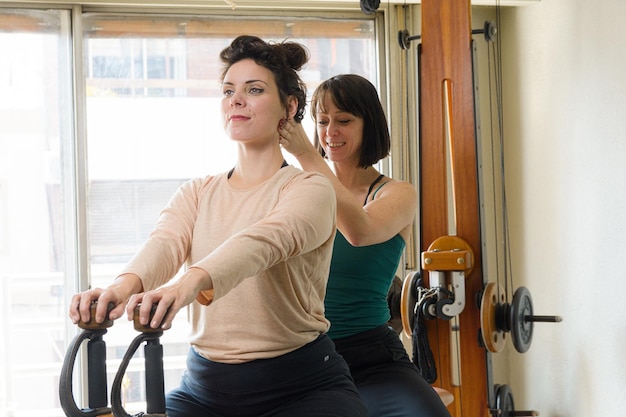 Instrutora de exercícios latina adulta corrigindo a postura espinhal de sua aluna sentada na máquina de exercícios em casa