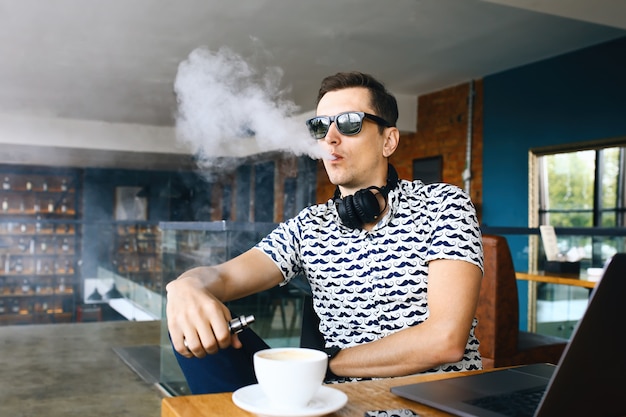 Foto insunglasse de homem jovem hippie bonito sentado no café com uma xícara de café, vaping e libera uma nuvem de vapor