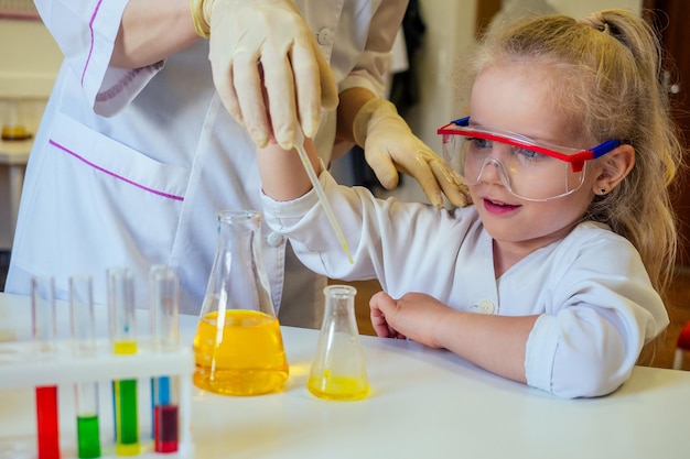 Inteligente colegiala rubia química en anteojos y bata médica blanca haciendo experimentos científicos química mezclando diferentes soluciones químicas en el aula de laboratorio