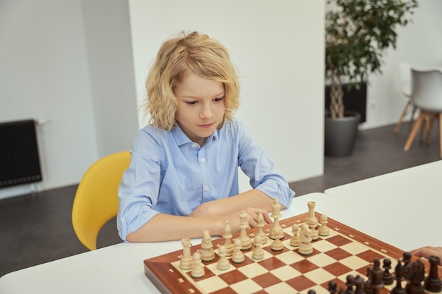 Intellektuelles Spiel intelligenter kaukasischer Junge im blauen Hemd, der im Klassenzimmer sitzt und Schach spielt