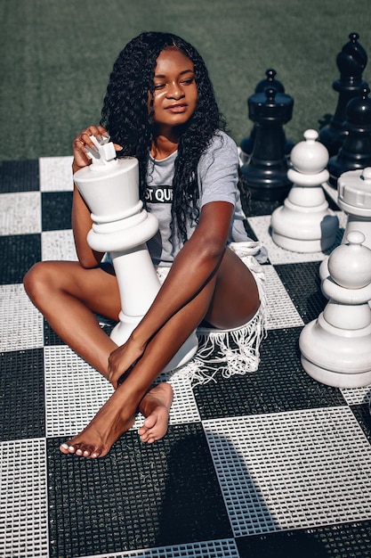 Intelligente afroamerikanische Frau, die Riesenschach spielt, während sie mit gekreuzten Beinen auf dem Brett sitzt; glamouröses intelligenzkonzept.