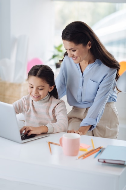 Intelligente Generation. Positives nettes kluges Mädchen, das am Tisch sitzt und auf dem Laptop tippt, während Zeit mit ihrer Mutter verbringt