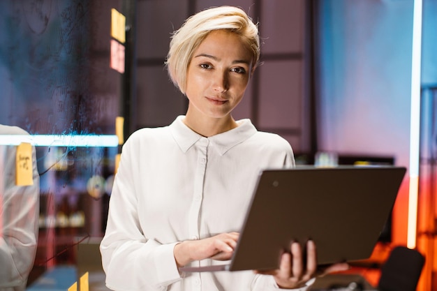 Intelligente Geschäftsfrau mit weißem Hemd, die auf einem Laptop tippt, während sie in der Nähe einer transparenten Glaswand steht