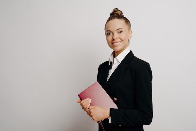 Intelligente, glückliche Geschäftsfrau hält ein rotes Notebook, lächelt zur Kamera und posiert isoliert vor einem hellen Hintergrund