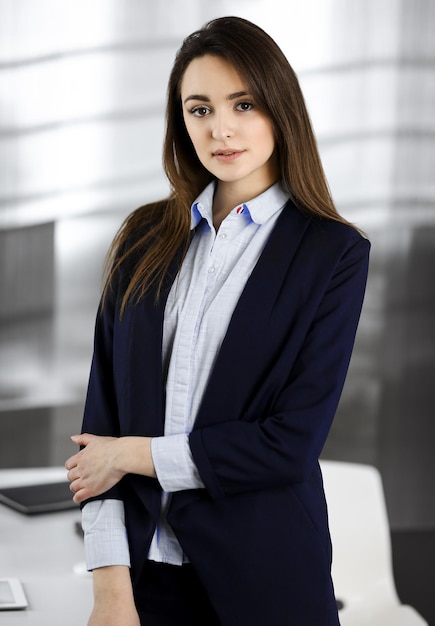 Intelligente junge Geschäftsfrau in einer blauen Jacke steht an ihrem Arbeitsplatz. Porträt eines Spezialisten in einem modernen Büro.