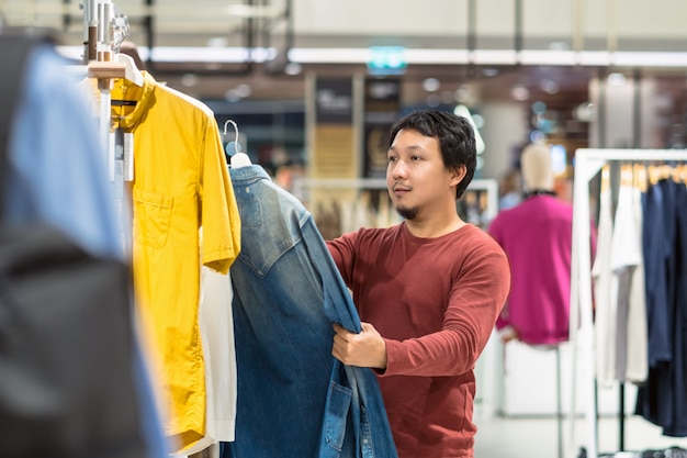 Intelligenter asiatischer Mann mit dem Bart, der Kleidung im Bekleidungsgeschäft im Einkaufszentrum wählt