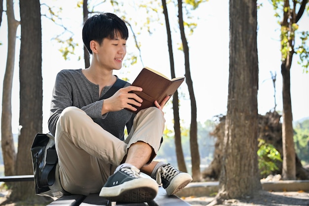 Intelligenter und chilliger junger asiatischer männlicher College-Student, der ein Buch liest, während er sich im Park entspannt