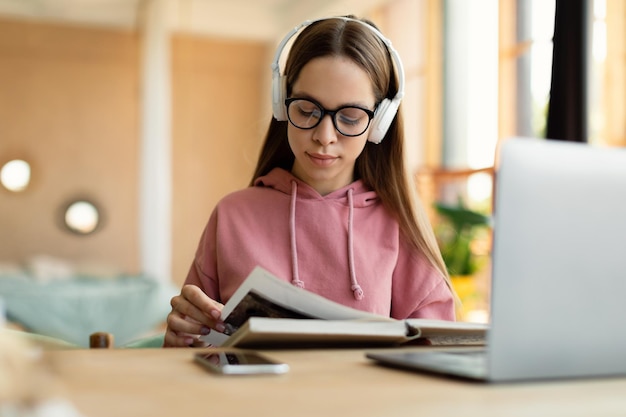 Intelligentes jugendlich Mädchen, das Lehrbuch liest, das mit Laptop am Tisch sitzt und Materialien lernt