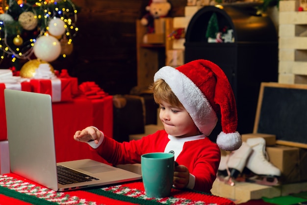 Intelligentes Surfen im Internet für Kleinkinder. Kleiner Junge Weihnachtsmütze und Kostüm. Jungenkind mit Laptop nahe Weihnachtsbaum. Kaufen Sie Weihnachtsgeschenke online. Weihnachtseinkaufskonzept. Geschenkservice. Santa kleiner Helfer.