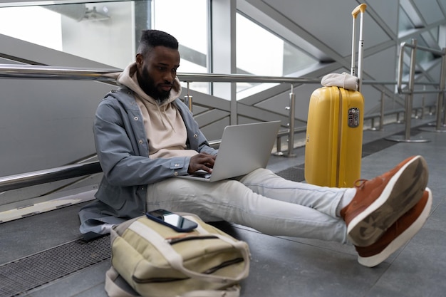 Intenção jovem negro com computador nas voltas sentado no chão do aeroporto reservando apartamento para viagem