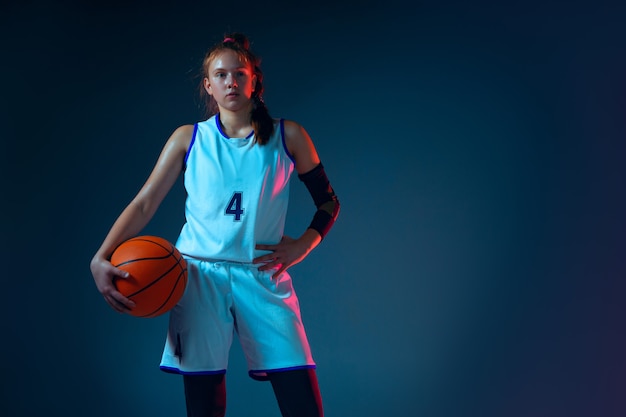 Intensidade. jovem jogadora de basquete feminino caucasiano sobre fundo azul studio em luz de néon, movimento e ação. conceito de esporte, movimento, energia e estilo de vida dinâmico e saudável. posando confiante.