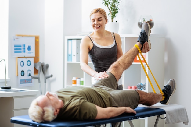 Intente más alto. Bonita mujer feliz sosteniendo la pierna de su paciente mientras lo ayuda a levantarla