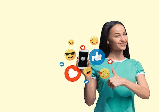 Interações de mídia social no celular. marketing digital na internet, conversando, comentando, curtindo. sorrisos e ícones acima da tela do smartphone, que segurando por uma jovem no fundo amarelo do estúdio.