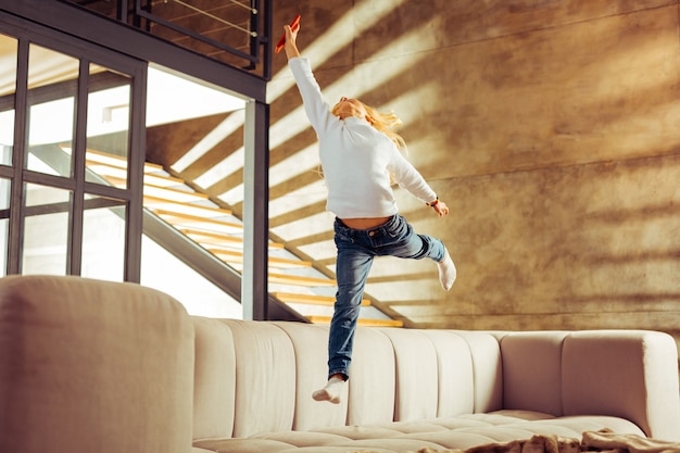 Interaktion zu Hause. Begeistertes blondes Kind hält das Gleichgewicht beim Springen auf dem Sofa balance