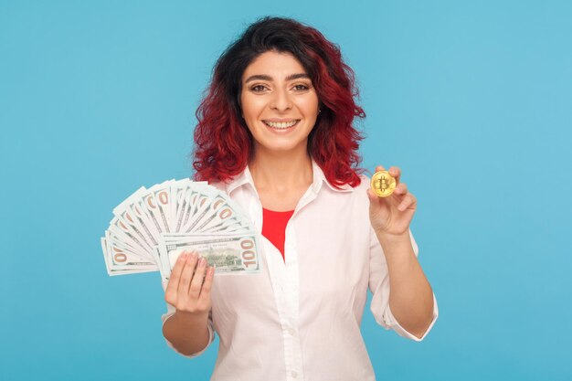 Intercambio de criptomonedas. Una mujer alegre y sonriente con el pelo rojo elegante sosteniendo billetes de bitcoin y dólares, dinero digital, comercio electrónico. Foto de estudio interior aislado sobre fondo azul.