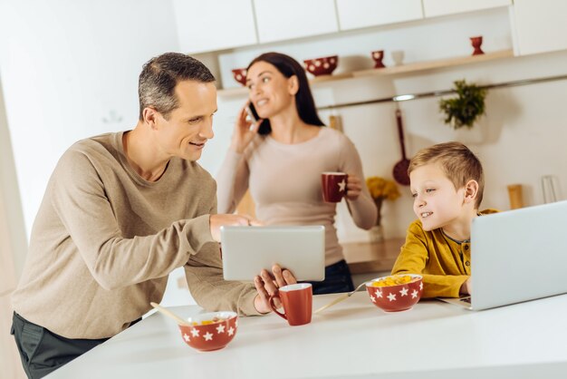 Interesante discusión. Agradable joven padre mostrando a su pequeño hijo una tableta con un video y discutiéndolo con el niño en lugar de desayunar